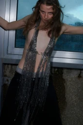 Model wearing a shimmering chainmail halter top and dark pants leaning against a large window.