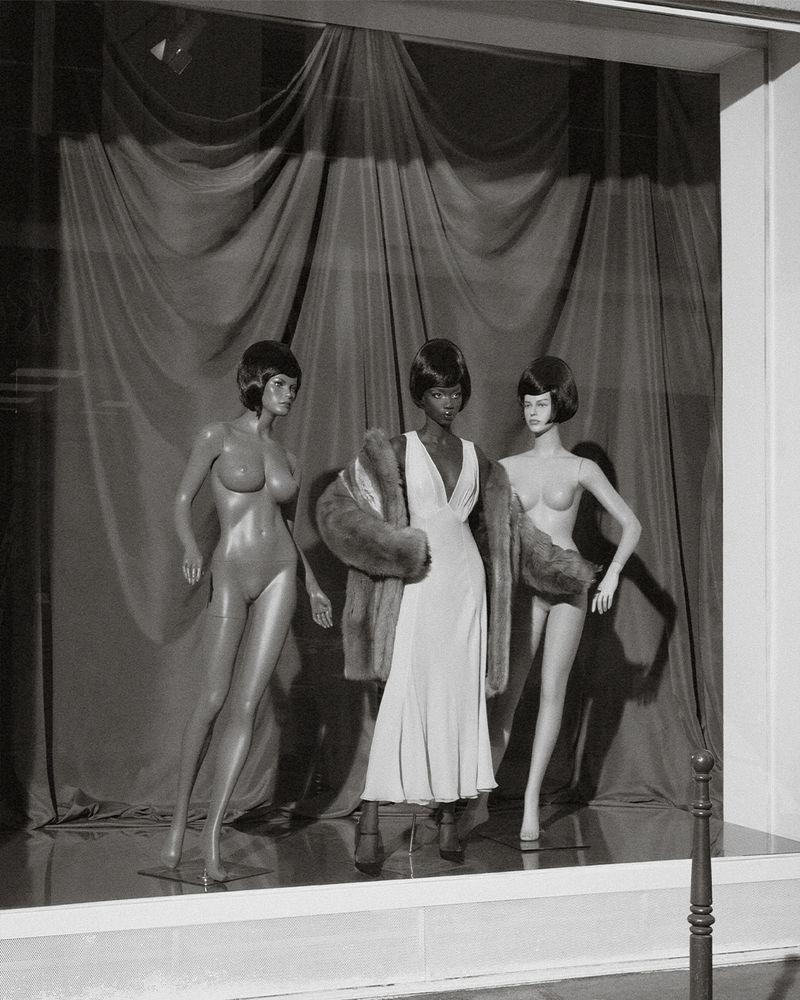 Monochrome shot of three mannequins posed in a store window wearing a dress and fur coat, displayed against a draped backdrop.