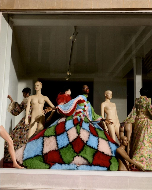 Group of mannequins dressed in colorful fashion, including a large patchwork dress, in an indoor, presumably retail or display setting.
