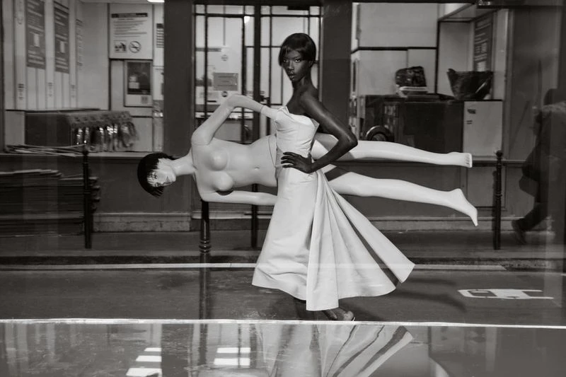 Black and white shot of a Black woman in a white dress with her hand on her hip, next to a mannequin in a store window.