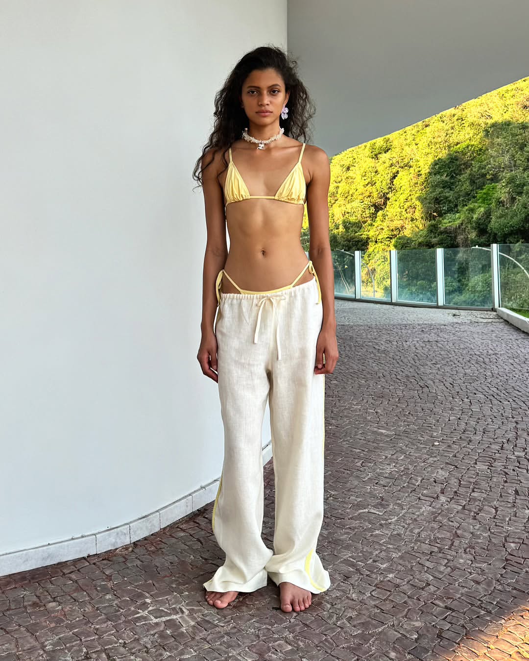 Woman in yellow bikini top and linen pants poses outdoors with a mountain backdrop on a paved walkway in natural light.