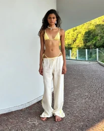Woman with long dark hair wearing a yellow bikini top and white drawstring trousers outdoors on a paved terrace.
