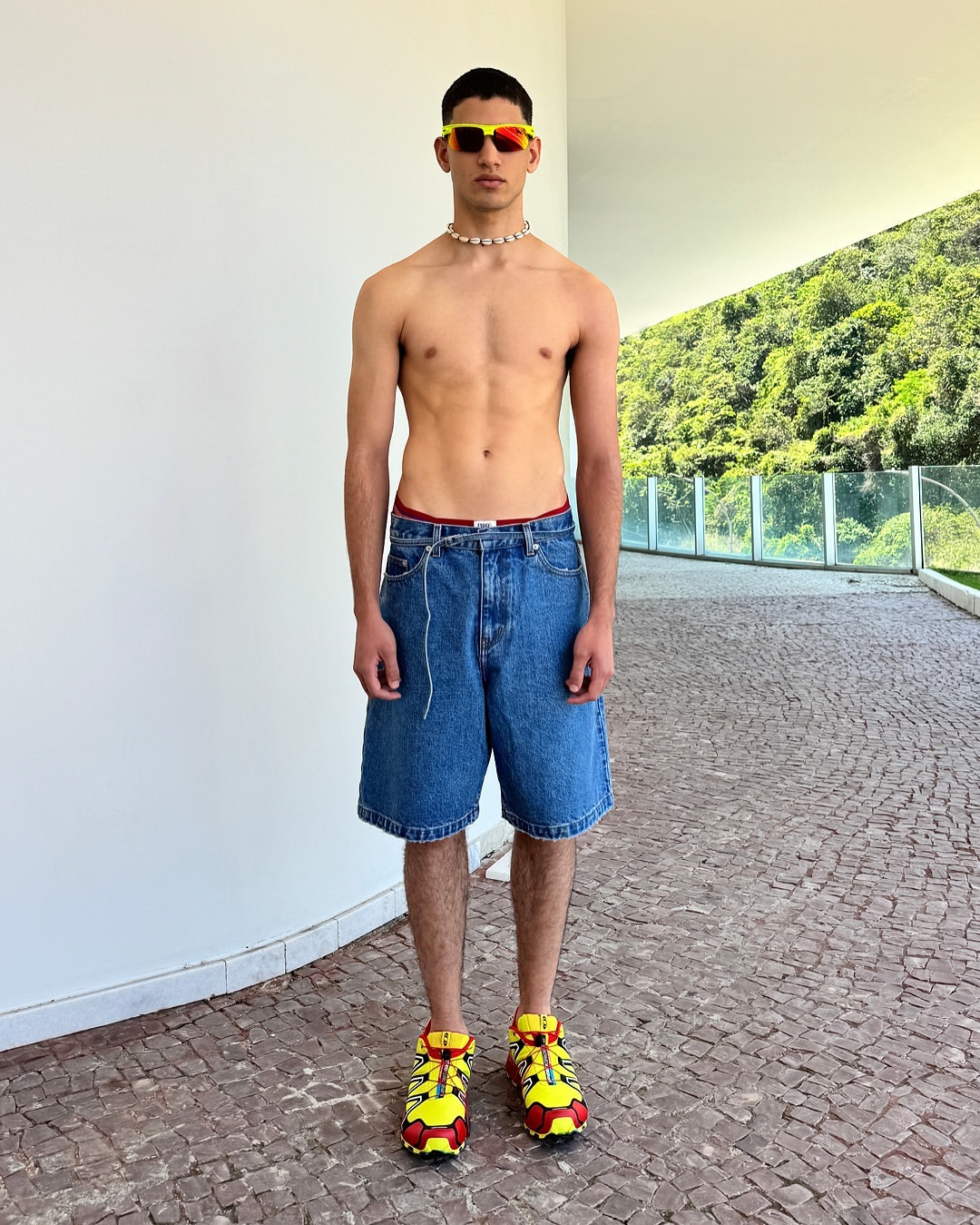 Full shot of a young adult man with an athletic build posing outdoors wearing sunglasses, necklace, denim shorts, and colorful sneakers.