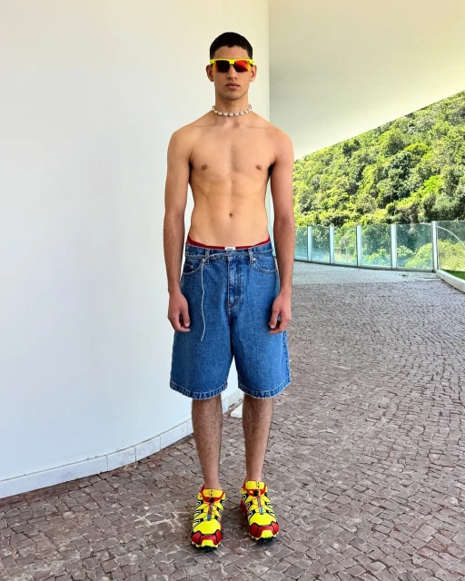 Full shot of a young adult man with an athletic build posing outdoors wearing sunglasses, necklace, denim shorts, and colorful sneakers.