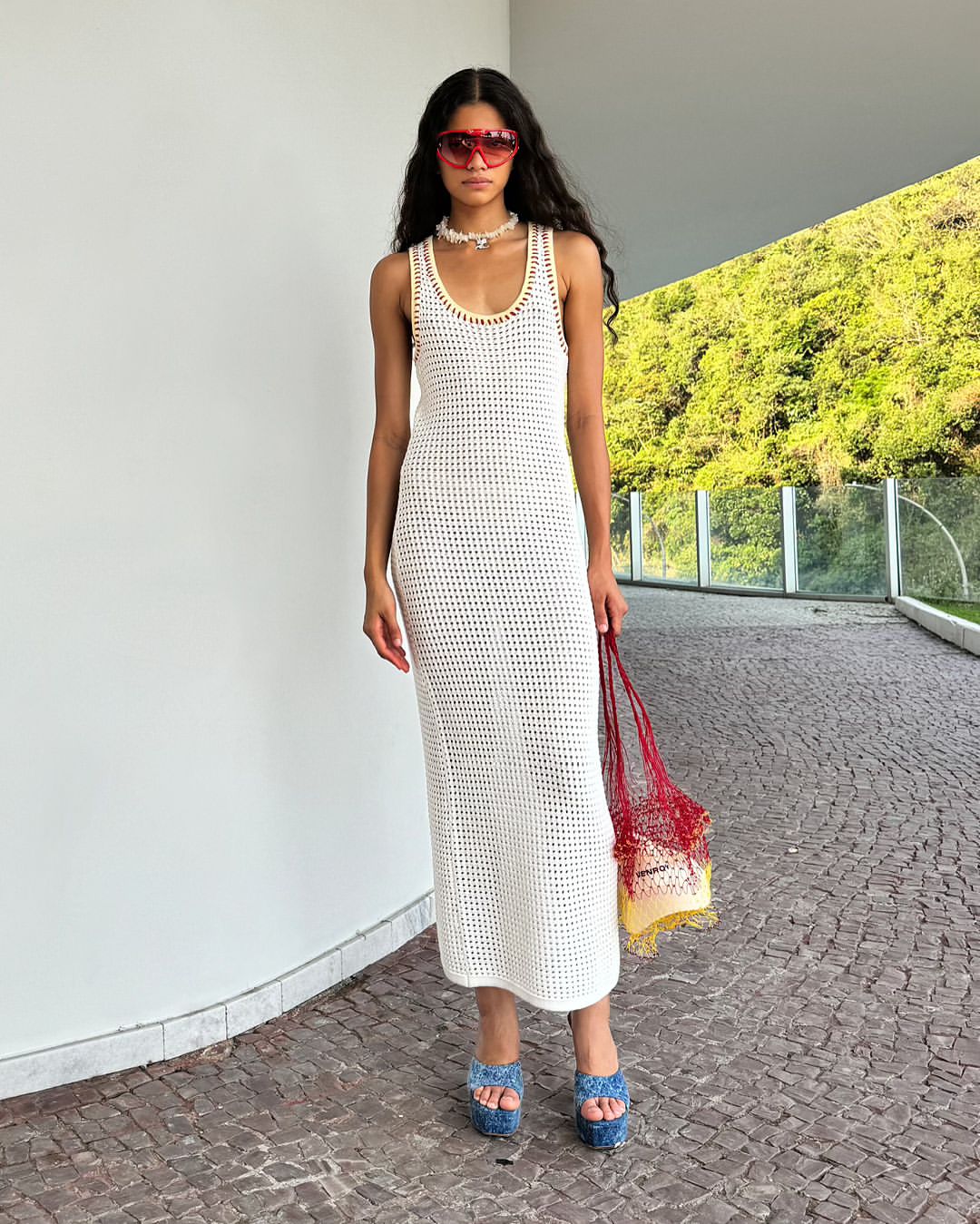 A woman stands on a paved area in a white dress, sunglasses, and blue platform sandals, holding a red net bag, showcasing a summer fashion style.