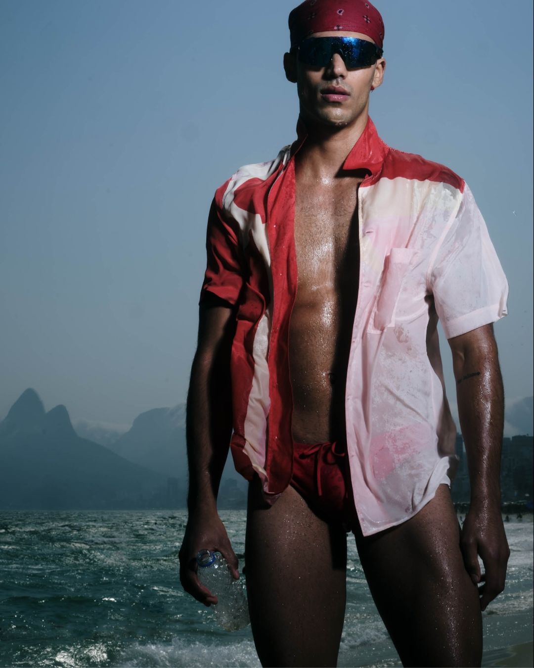 Athletic man poses by the ocean wearing an open shirt, swim trunks, bandana, and sunglasses, holding a water bottle with mountains in background.