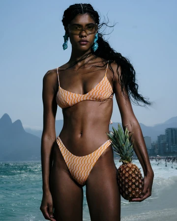 Woman in patterned bikini holds pineapple on beach with mountain background. Fashion editorial photo in Rio de Janeiro, Brazil.