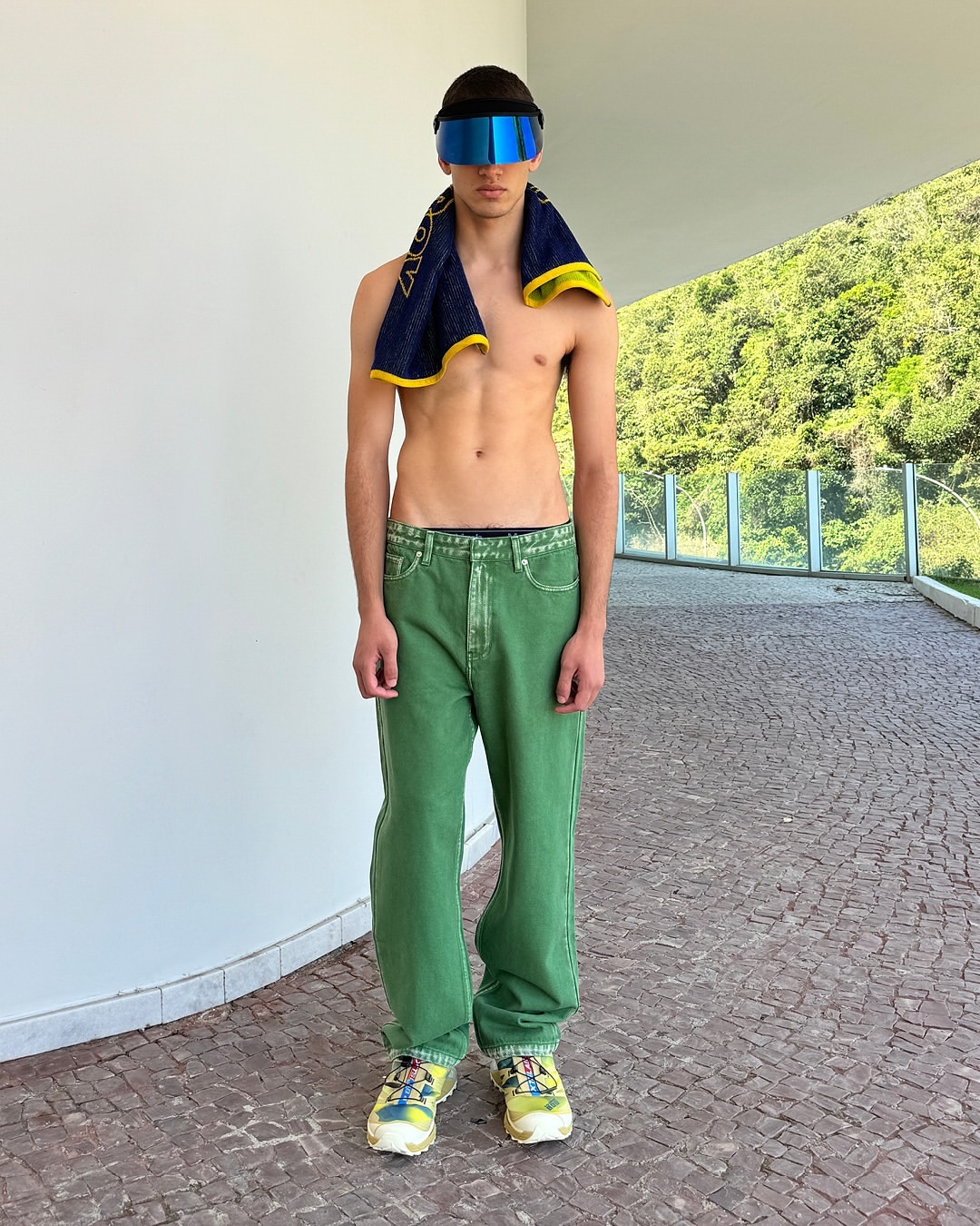 Athletic, shirtless man stands with towel and futuristic visor against a modern wall, with green trees visible in background.