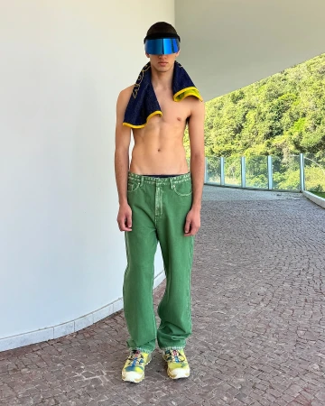 Athletic, shirtless man stands with towel and futuristic visor against a modern wall, with green trees visible in background.