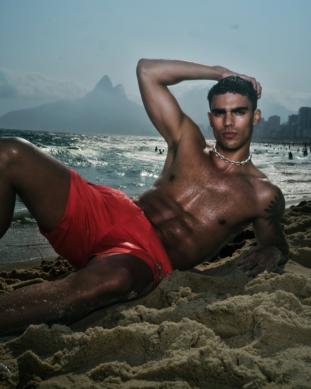 Man in red swim trunks poses on a sandy beach with ocean waves and Rio de Janeiro mountains in the background on a sunny day.