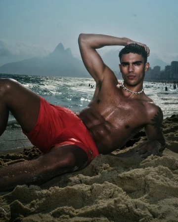 Man in red swim trunks poses on a sandy beach with ocean waves and Rio de Janeiro mountains in the background on a sunny day.