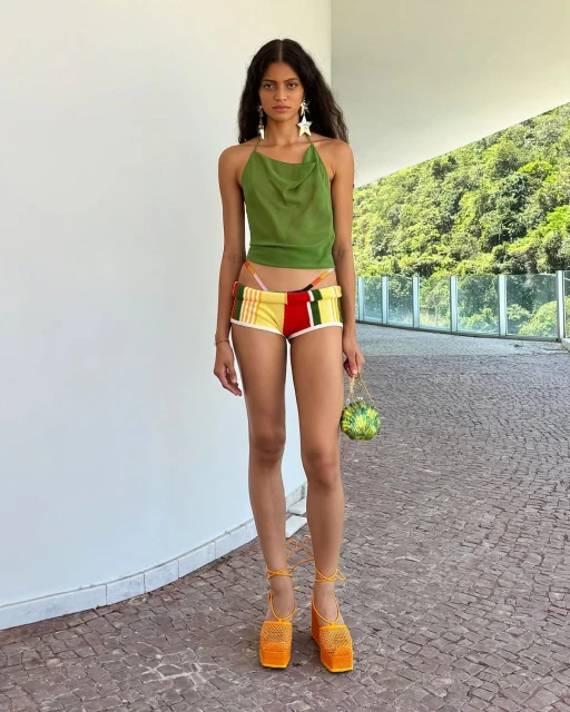 Full shot of a young woman in a green top and multicolor shorts, standing outside on a brick surface. She is holding a round bag.