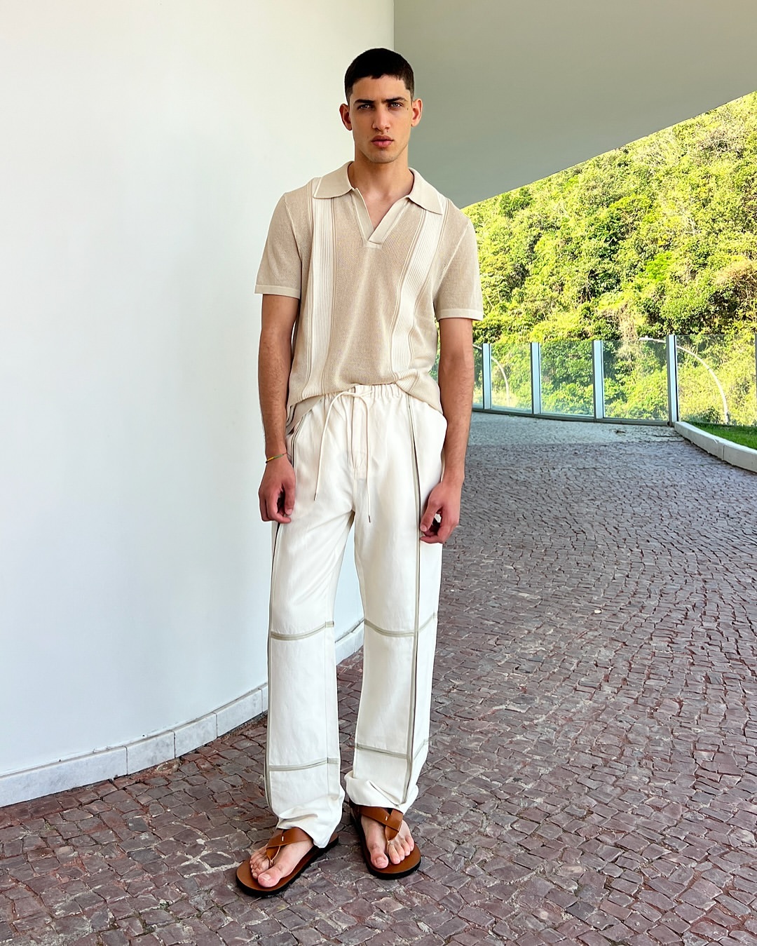Man in a casual outfit, wearing a beige knit polo shirt and white drawstring pants, standing outside in front of green foliage.