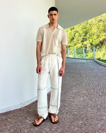 Man in a casual outfit, wearing a beige knit polo shirt and white drawstring pants, standing outside in front of green foliage.