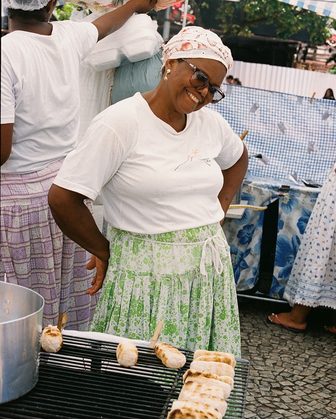 Smiling Afro-Brazilian woman cooking acarajé on a grill at an outdoor food market wearing a headscarf, white t-shirt and green skirt.