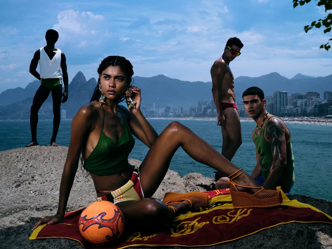 Four people on a rocky outcrop with Rio de Janeiro skyline in the background. One woman is seated with a soccer ball.