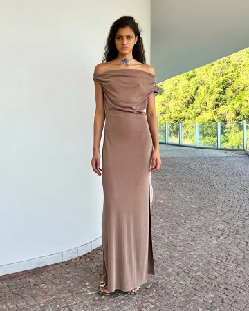 Elegant adult woman in a long, mocha-colored dress poses outside on a stone patio with a background of green trees and white walls.
