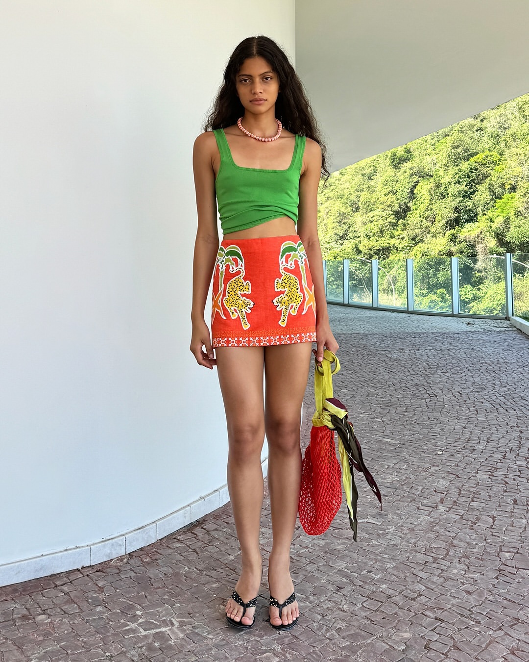 A full-length portrait of a young woman in an orange skirt with an animal print, a green tank top, black sandals, and a yellow bag.