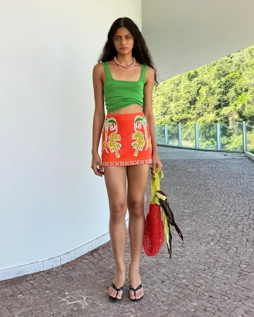 A full-length portrait of a young woman in an orange skirt with an animal print, a green tank top, black sandals, and a yellow bag.
