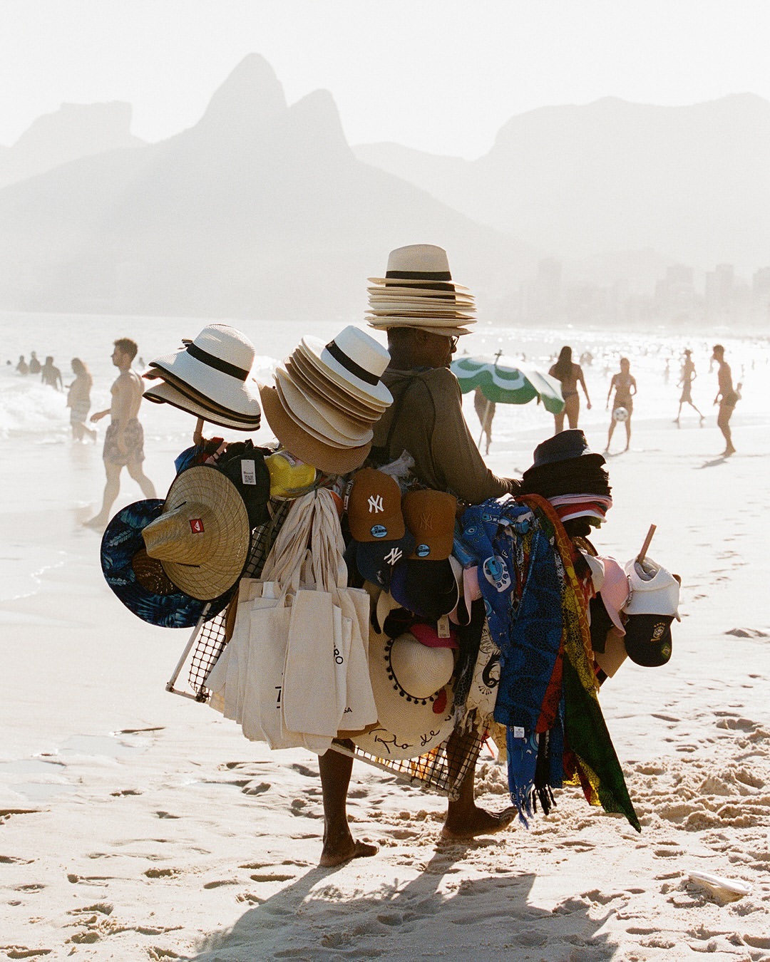 A street vendor on a sandy beach carries a stack of hats and bags, with people in the ocean and a mountain range in the background.