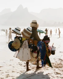 Vendor carrying a large rack of straw hats and beach bags along a sunny coastal beach with mountains in the background
