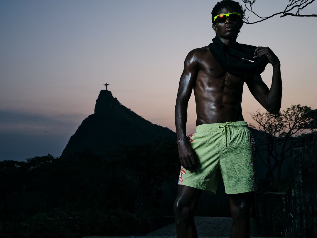 Dark-skinned adult male wearing sunglasses and lime shorts stands outdoors with a scarf and mountain backdrop against the sunset.