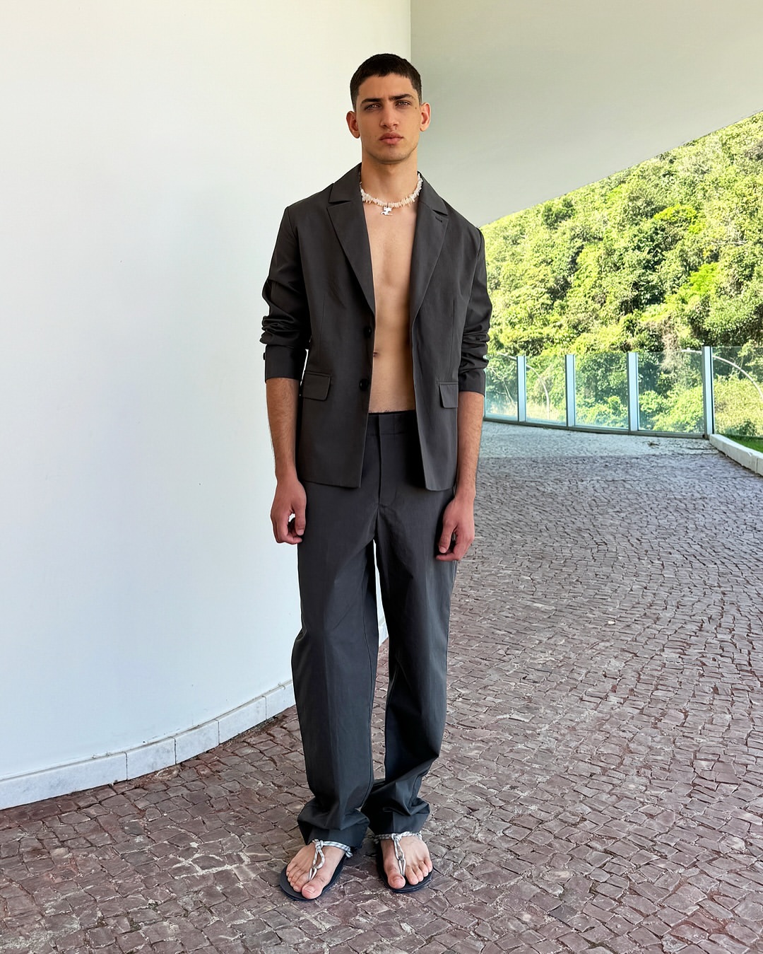 An adult male in an open suit jacket and trousers, paired with flip-flops and a beaded necklace, poses in front of a paved walkway and greenery.