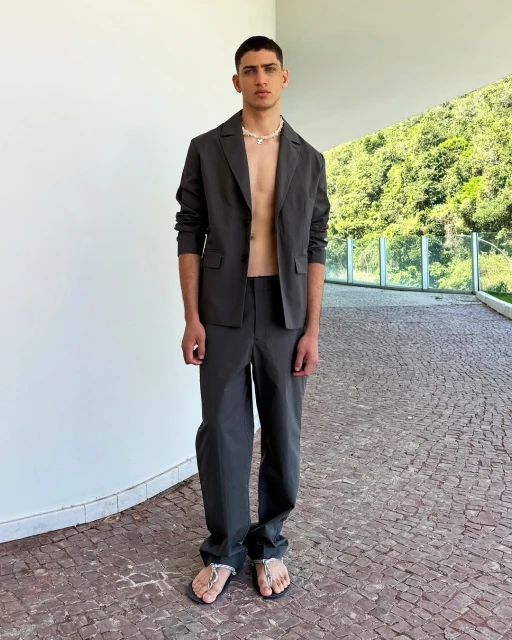 An adult male in an open suit jacket and trousers, paired with flip-flops and a beaded necklace, poses in front of a paved walkway and greenery.