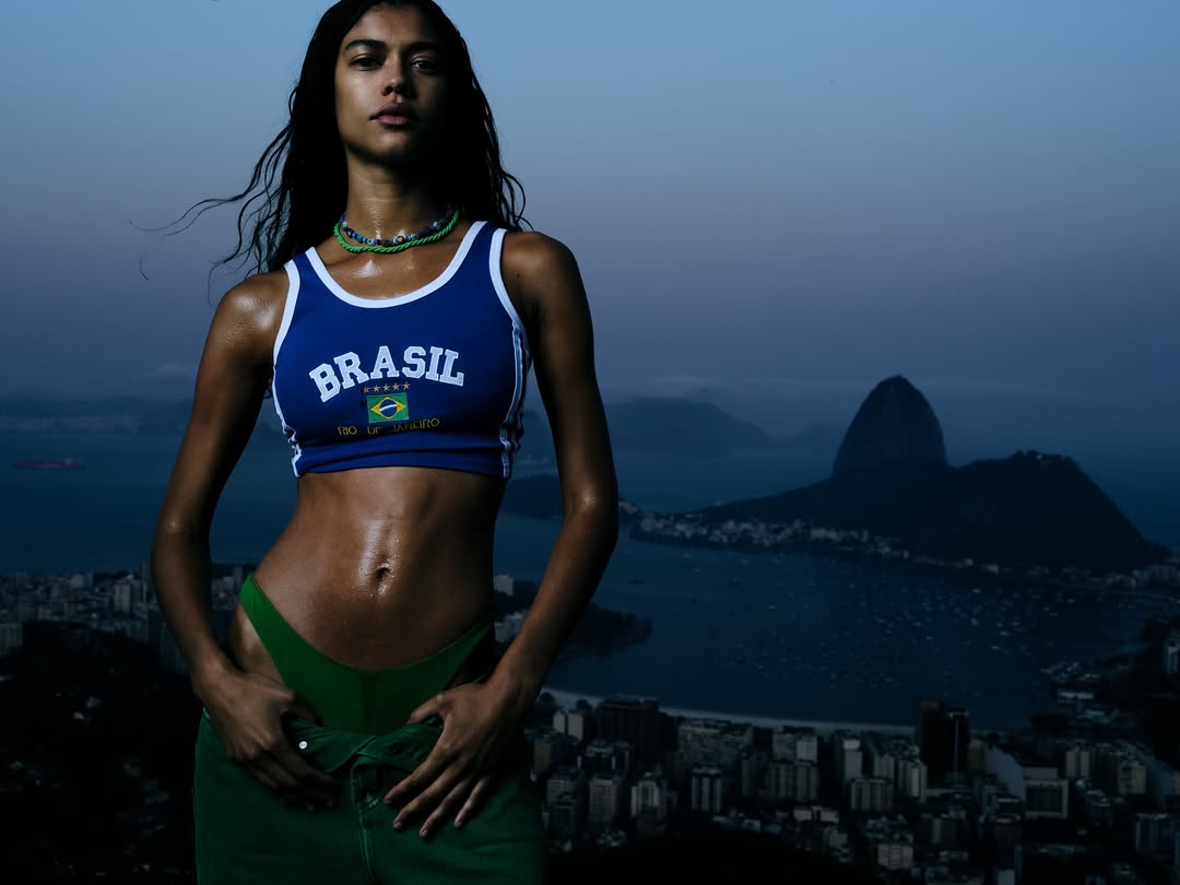 Woman in athletic top with 'Brasil' lettering poses with Rio de Janeiro cityscape and Sugarloaf Mountain in background at dusk.
