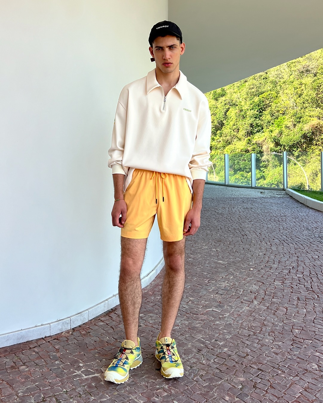 Full-length portrait of a young adult male in a casual outfit, featuring a long-sleeve shirt, yellow shorts, and athletic shoes, outdoors.
