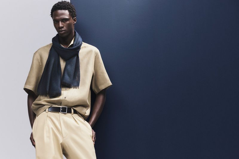 Elegant adult male model in casual tan shirt, draped dark scarf, and tan trousers posing in a studio against a split blue and white backdrop.