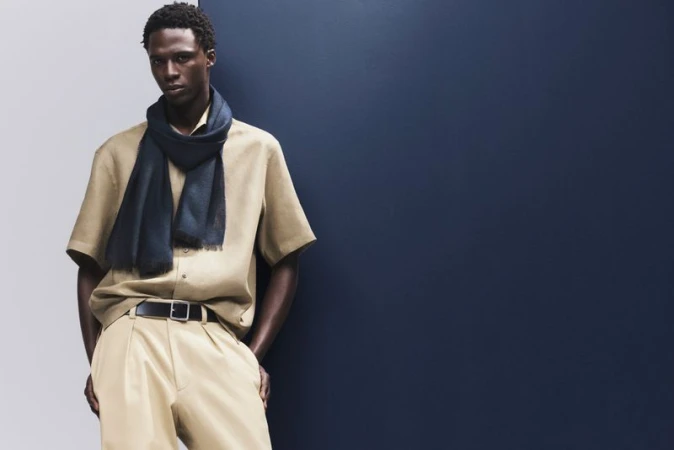 Elegant adult male model in casual tan shirt, draped dark scarf, and tan trousers posing in a studio against a split blue and white backdrop.