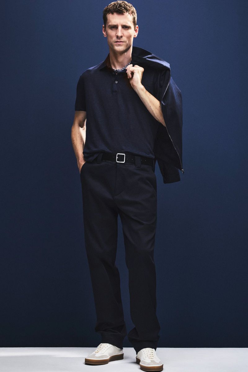 A full shot shows a man in navy polo shirt and trousers, with jacket over shoulder, against a dark blue backdrop in studio lighting.