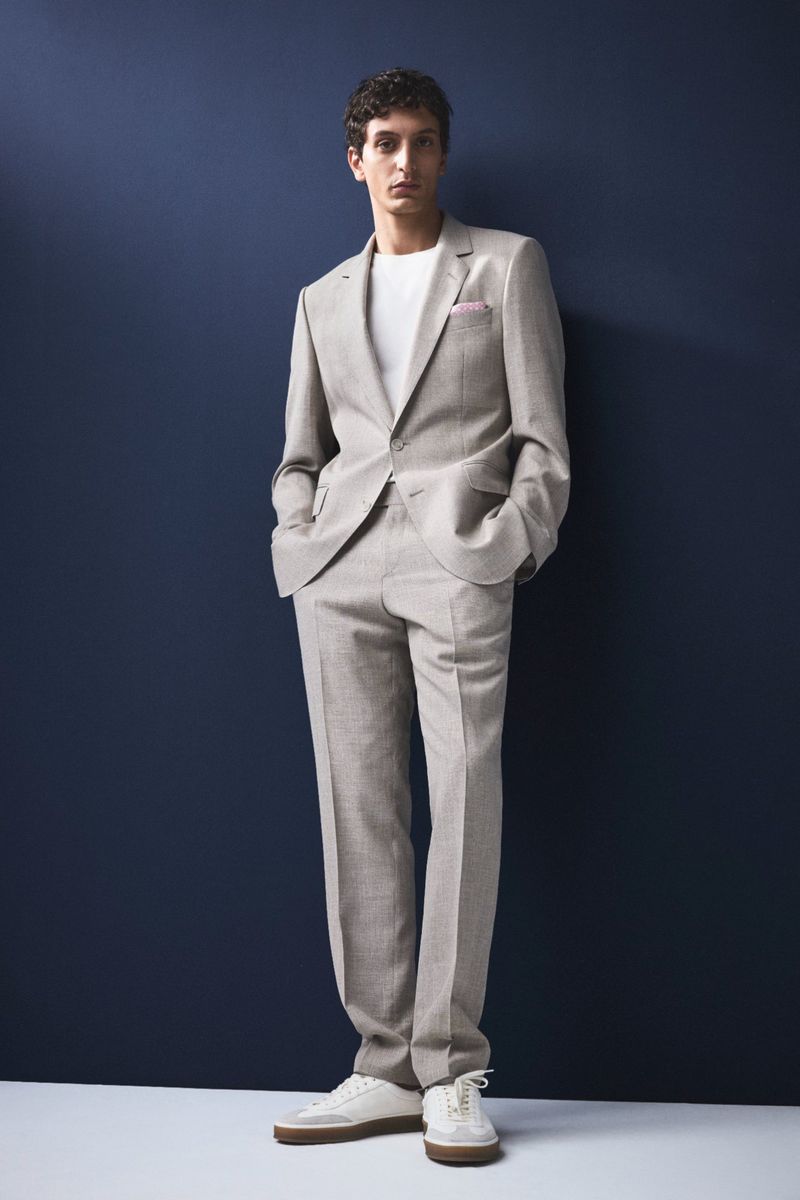 Adult male posing in studio wearing a light tan suit with white trainers against a dark blue background in a full studio shot.