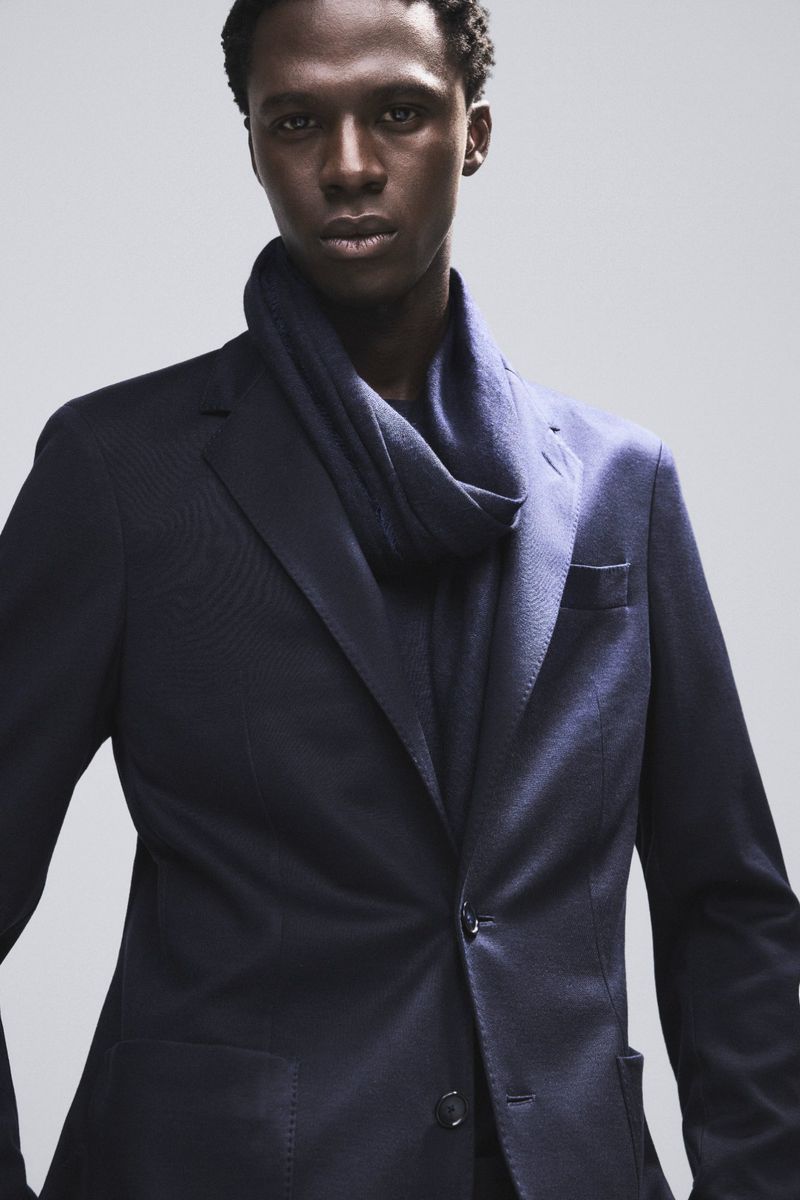 Elegant dark-skinned man in a navy blazer and scarf stands in a photo studio, exuding sophistication and style in a medium studio shot.