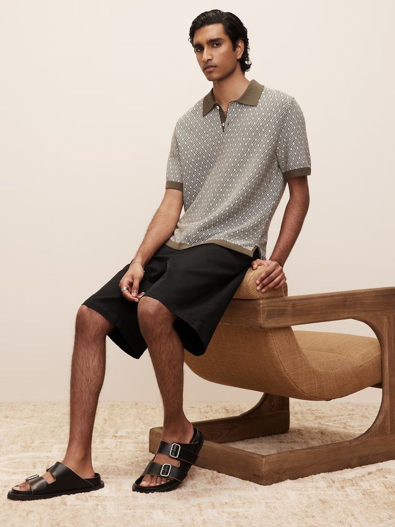 A dark-haired adult male sits on a chair wearing a knit polo shirt with shorts and sandals, photographed in a light indoor setting.