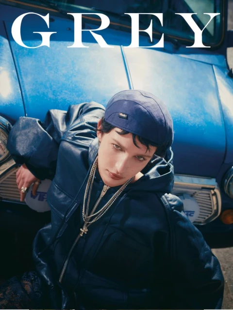 Fashion shot of a dark-haired model in a black jacket, cap, and chain necklace, leaning against a blue car outdoors.