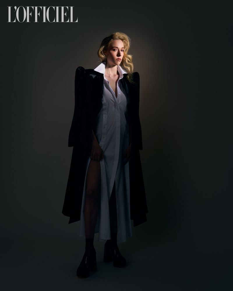 A full-length portrait showcases a woman with wavy blonde hair, wearing a white dress, black coat, and heels against a dark backdrop.