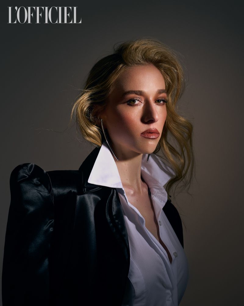 Blonde woman in a fashion portrait, wearing a white collared shirt and black jacket against a grey backdrop with soft studio lighting.