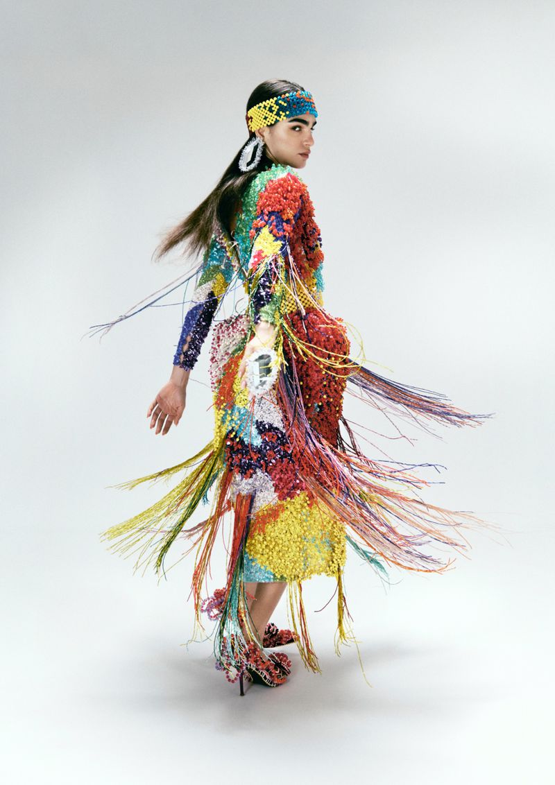A female fashion model in a colorful beaded dress and headdress poses in a studio with a white background, turned to look at the camera.