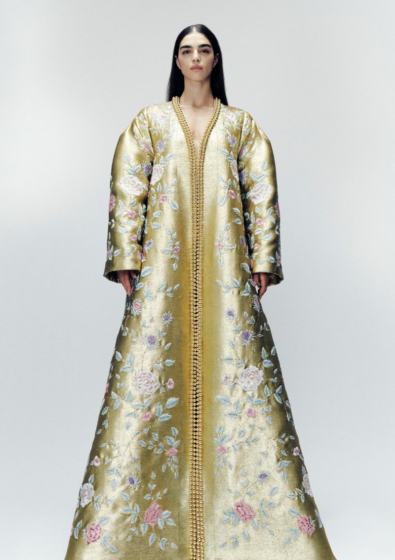 Full-length portrait of a woman in a gold gown with floral embroidery standing in a white photo studio. The garment is floor-length.