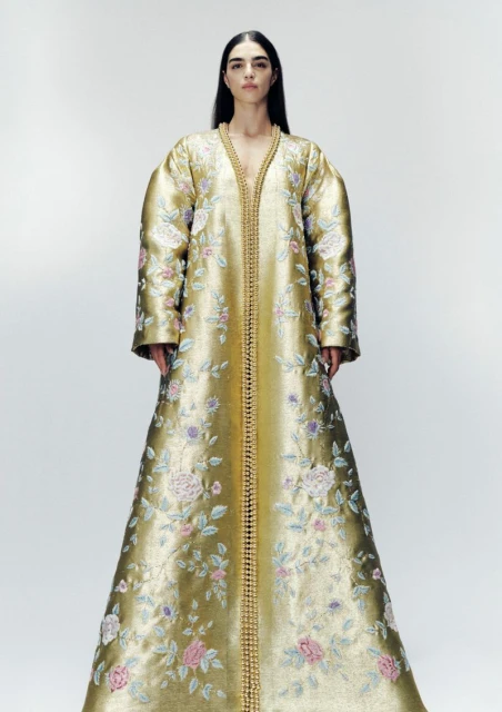 Full-length portrait of a woman in a gold gown with floral embroidery standing in a white photo studio. The garment is floor-length.