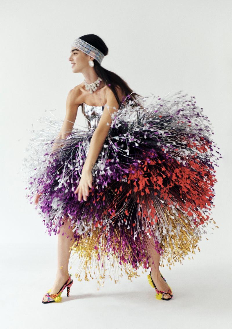 A slender adult woman smiles in profile, modeling a festive couture dress made from metallic, colorful tinsel in a studio setting.