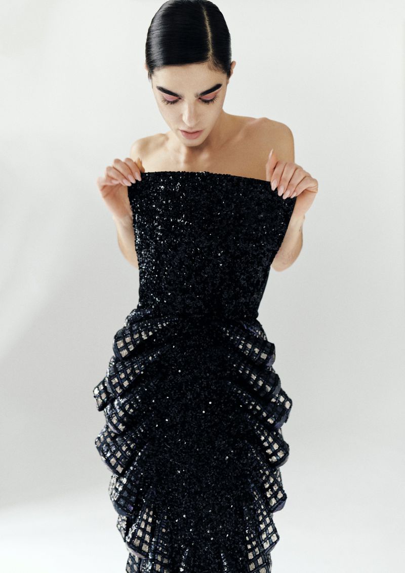 A woman in a strapless black sequin gown stands in a photo studio, looking down at the dress. Fashion and beauty concept.