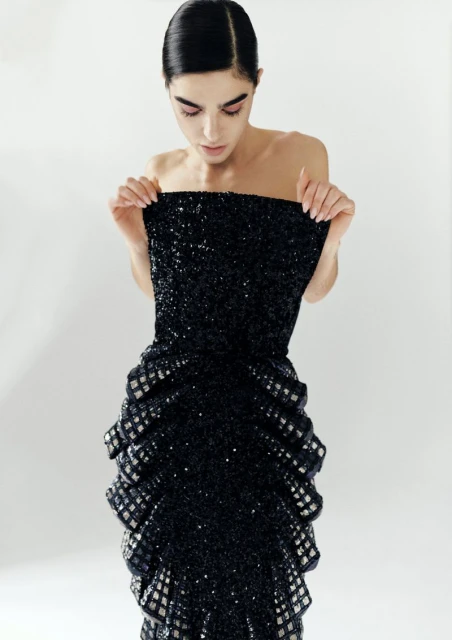 A woman in a strapless black sequin gown stands in a photo studio, looking down at the dress. Fashion and beauty concept.