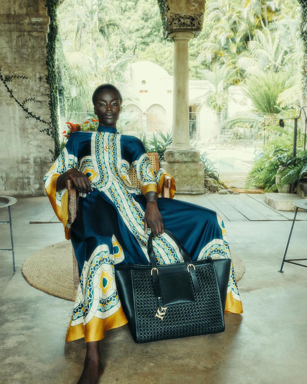 Woman in patterned caftan poses with a black leather tote bag on a veranda overlooking a green landscape, captured in soft, diffused light.