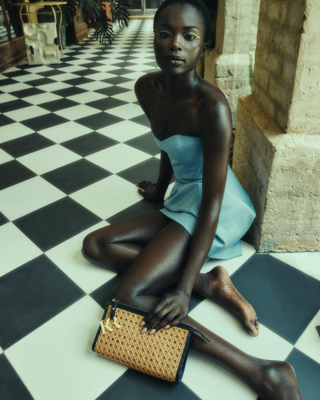 A Black woman with gold eyeliner and a light blue dress poses on a black and white tiled floor, holding a wicker purse.