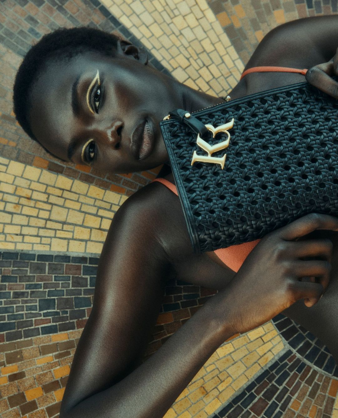 Attractive adult woman laying down on patterned surface holding a black clutch purse with gold colored logo, wearing orange top.