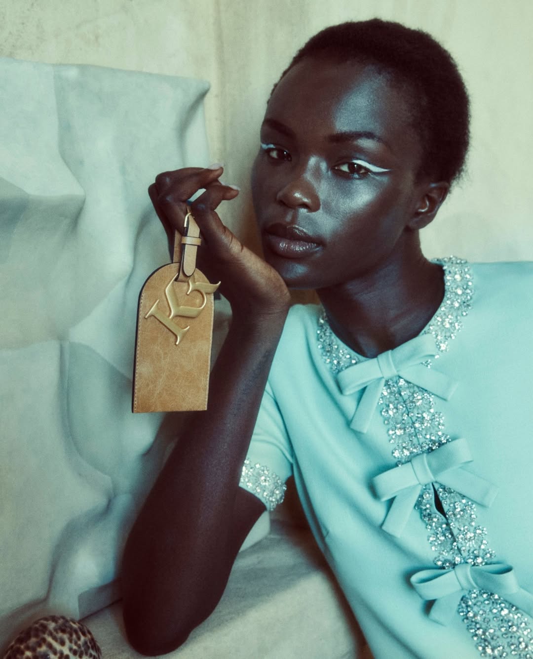 Elegant dark-skinned model showcasing a light blue dress with bow accents, holding a 'LV' branded luggage tag in a sophisticated studio setting.