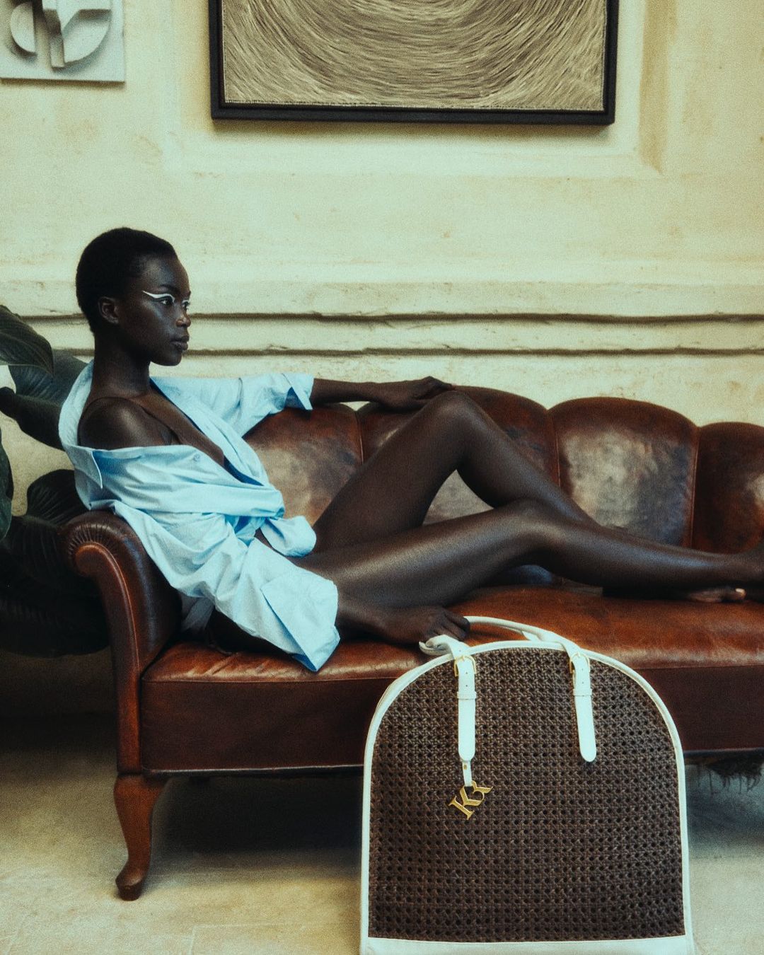Elegant black woman in a light blue shirt lounging on a leather couch indoors with a wicker tote bag, art on the wall.